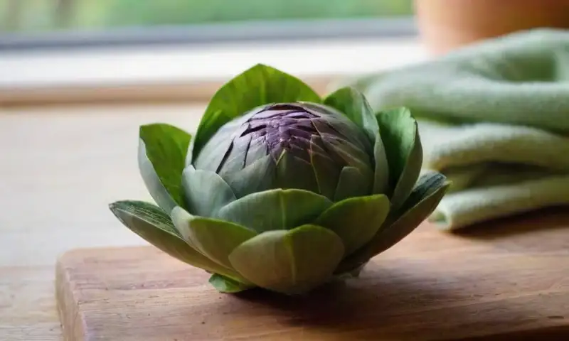 Una alcachofa verde brillante se destaca sobre una tabla de madera rústica, rodeada de utensilios de cocina y un ambiente acogedor