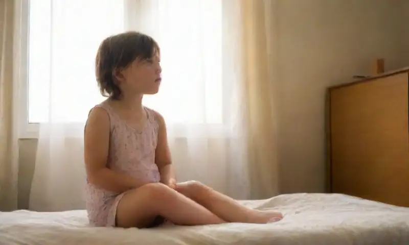 Un niño cansado y vulnerable se sienta en una cama deshecha en un cuarto tenue, rodeado de luz suave, con dibujos coloridos en las paredes que contrastan con su fragilidad