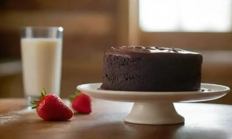 Una mesa de madera rústica recibe luz cálida, rodeada de leche cremosa, un pastel de chocolate, fresas y crema batida, creando un ambiente acogedor
