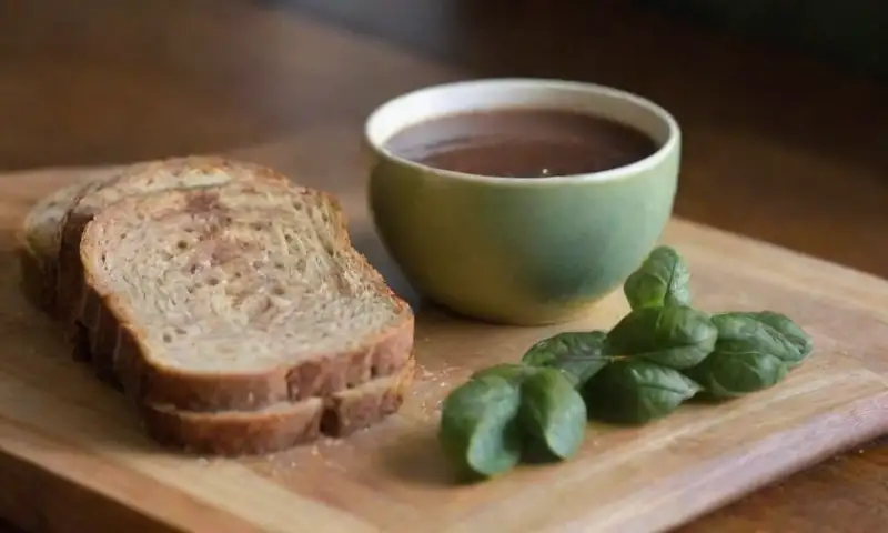 Una mesa de madera presenta un acogedor fondo con una taza de chocolate caliente y diversos alimentos coloridos y texturizados, iluminados suavemente por la luz que entra por la ventana