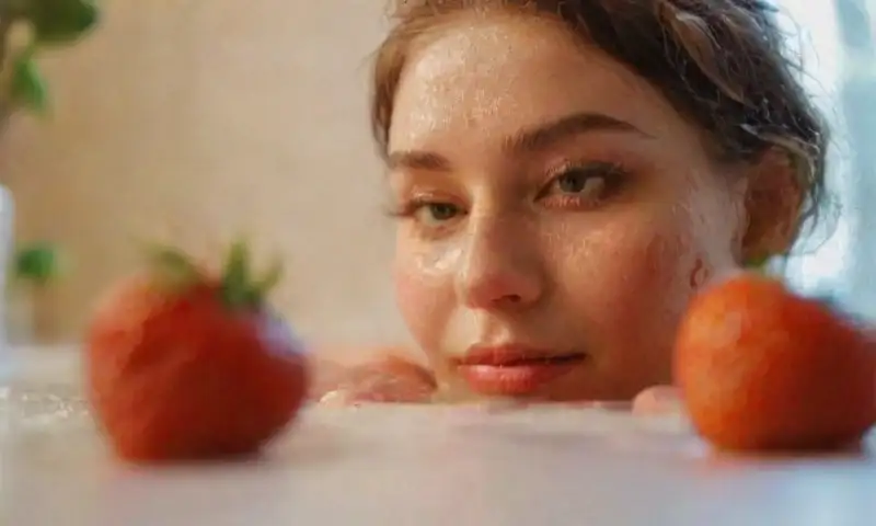 Una joven con piel marrón clara y fresas rojas en las mejillas está en un baño iluminado suavemente, con un ambiente tranquilo