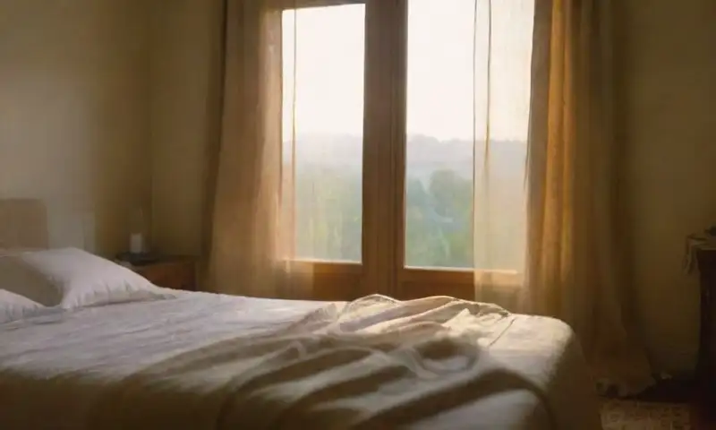 Un dormitorio tenue al amanecer con luz dorada, una cama deshecha, un vaso de agua en la mesa y una mano emergiendo bajo la manta, todo envuelto en una atmósfera de calma y calidez