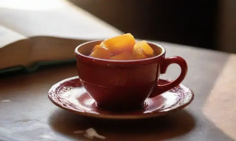 Una cálida mesa de madera ilumina una taza de té rojo sobre un plato de porcelana, acompañada de un libro y frutas frescas, creando un ambiente acogedor