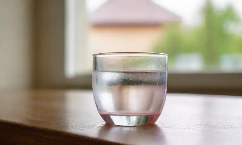 Un vaso de agua brilla sobre una mesa de madera, junto a una píldora y un pañuelo arrugado, creando una escena contemplativa iluminada por luz natural